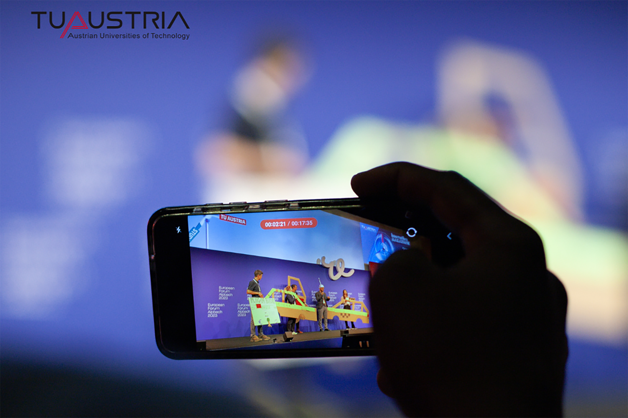 Hintergrund verschwommen, Vordergrund Hand inschwarz mit Handy, das Geschehen auf einer Bühne filmt