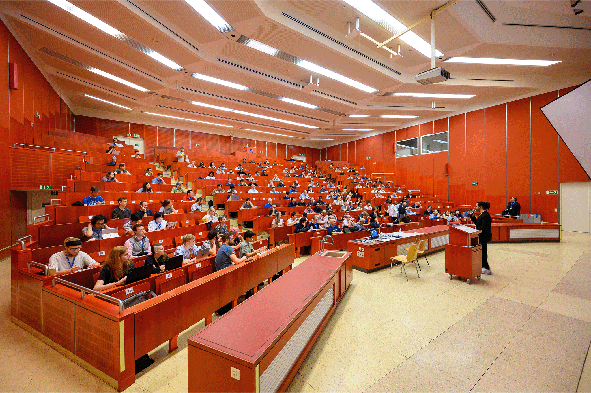 Hörsaal mit roten Wänden und Publikum mit vortragender Person