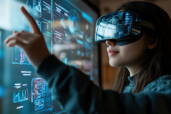 Junge Frau hat VR-Brille auf, auf der sich die Diagramme spiegeln, die sie auf einem Bildschirm ansieht. 