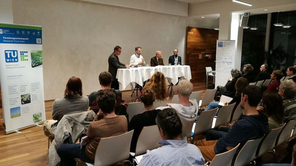 Zentral sind der Moderator und das Podium zu sehen. Davor sitzt das Publikum. Links steht ein Roll-Up des Forschungsschwerpunktes Energie und Umwelt.