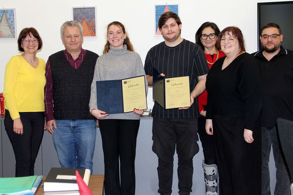 Gruppenfoto mit sieben Personen. In der Mitte die beiden Lehrlinge, die ihre geöffneten Urkunden in Händen halten.