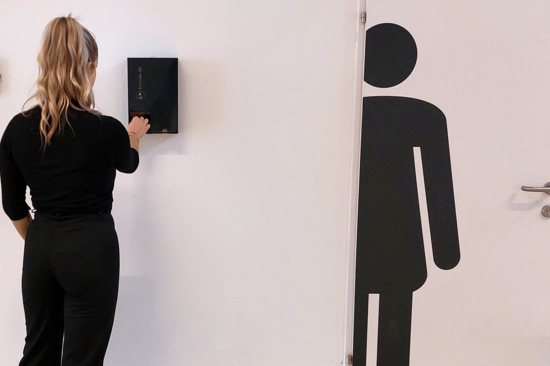 Schwarz gekleidete Person mit langen, zum Pferdeschwanz gebundenen blonden Haaren, die aus einem dunklen Spender ein Tampon holt. Rechts von ihr Toilettentür mit schwarzer, symbolischer Frauenfigur.