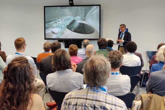 Menschen sitzend mit Rücken zur Kamera, vorne großer Bildschirm rechts daneben, Mann in Anzug