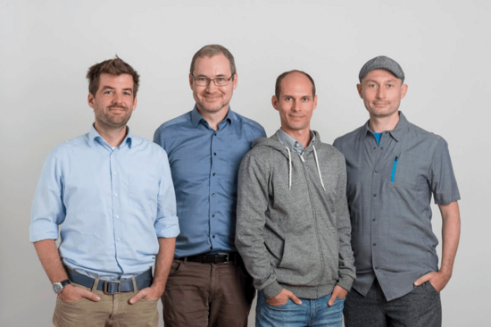 Vier Personen mit grauer und blauer Oberbekleidung, Hintergrund hellgrau. Drei haben ihre Hände in den Hosentaschen.