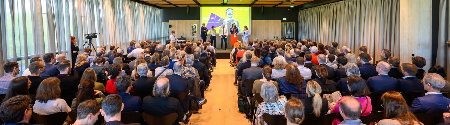 Saalaufnahme von der fuTUre fit Kick-Off Veranstaltung. Mehrere Menschen sitzen in Sesselreihen, auf der Bühne steht das Rektoratsteam