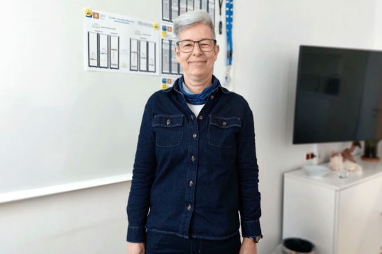 Christiane Philipp stehnend, mit dunkelblauem Hemd, Kurzhaarschnitt und Brille, grau-weiße Haare