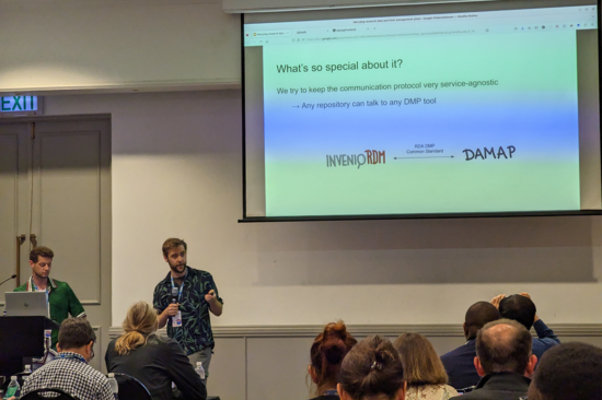 The photo shows the two speakers and a screen with their presentation plus some audience members in the foreground.