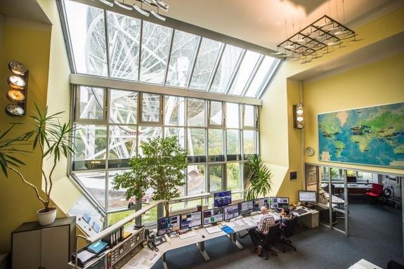 Photo from the control room of the radio telescope in the radio observatory Effelsberg. In the large room two employees are sitting in front of a row of screens. Through the large window front one can see the radio telescope.