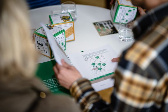 Zwei Personen lehnen sich über einen Tisch, auf dem sich Pappwürfel, eine Broschüre und Gläser befinden