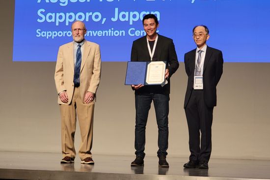 Gruppenfoto auf Bühne: In der Mitte der Preisträger Matthias Ikeda, Naoto Nagaosa, Chair der Commission C5: Low Temperature Physics der IUPAP, Matthias Ikeda (rechts), William P. Halperin, Vorgaenger von Naoto Nagaosa in dieser Rolle (links).