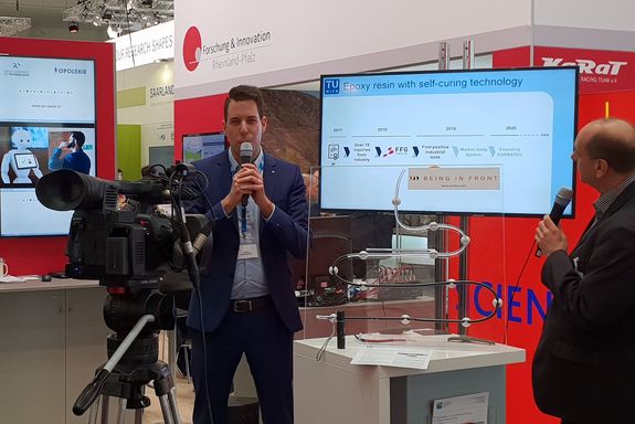 Daniel Grunenberg (TU Wien) explains the "Epoxy resin with self-curing technology" with a microphone in hands, infront of a camera and an audience at the "Science Square".