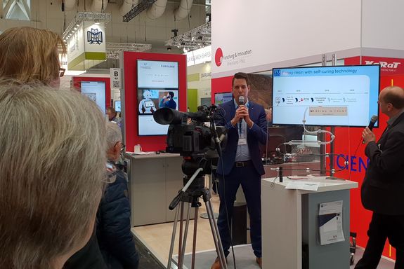 Daniel Grunenberg (TU Wien) explains the "Epoxy resin with self-curing technology" with a microphone in hands, infront of a camera and an audience at the "Science Square".