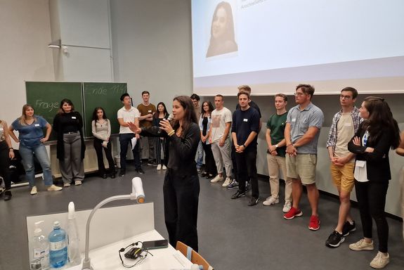Bühne mit Studierenden stehend im Halbkreis, davor eine Sprecherin mit Mikrofon
