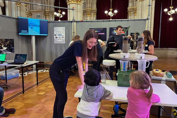 Zwei Kinder betrachten Experiment, während junge Forscherin dieses erklärt. 