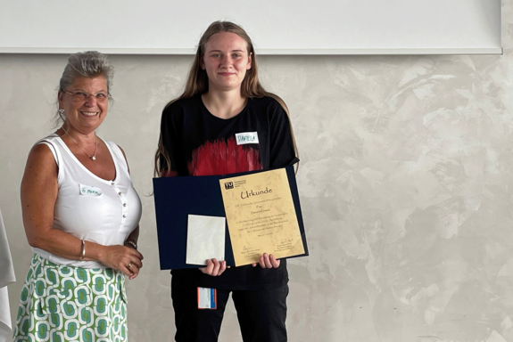 Manuela Marik, im weißen ärmellosen T-Shirt steht neben Daniela Gremel, in schwarzer Hose und schwarz-rotem T-Shirt. Sie hält eine Urkunde. Beide blicken direkt in die Kamera.