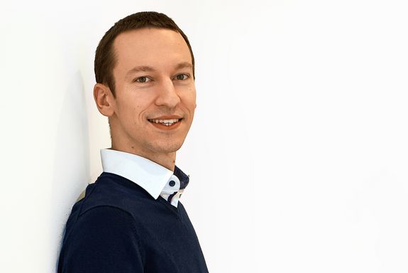 Portraitfoto von Bernhard Brenner, der an einer weißen Wand lehnt. Er trägt einen dunklen Pullover, darunter ein weißes Hemd, dessen Kragen man über dem Pullover sieht.