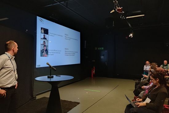 Das Veranstaltungsfoto zeigt Tomasz Miksa als Vortragenden, den Präsentationsbildshchirm inklusive Online-Teilnehmer_innen sowie die erste Reihe des Publikums.