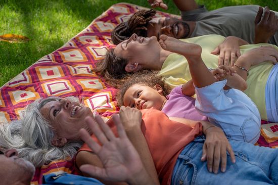 A diverse family (grandparents, parents, and child) are lying together on a picnic blanket and laughing.