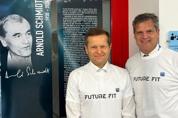 Foto mit einem Roll-Up auf dem Arnold Schmidt abgebildet ist, daneben Ferenc Krausz und TUW-Rektor Jens Schneider, die beide ein weißes T-Shirt mit der Aufschrift "Future Fit" tragen.