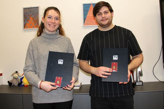 Zwei Personen, die eine Urkunde und davor einen kleinen Goldbarren halten