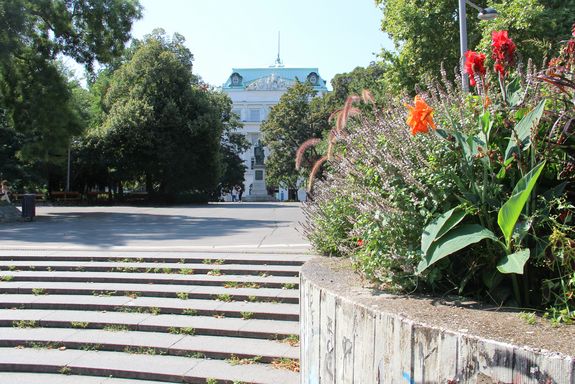 Sommerliche Ansicht auf das TU Wien Hauptgebäude, im Vordergrund rechts ein Beet mit roten Blumen, Lavendel und Ähren