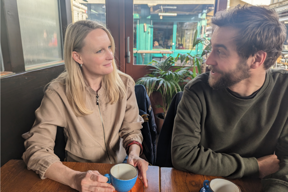 This photo shows Doris Matisovits and Maximilian Moser sitting next to each other at a table and looking at each other. On the table in front of them are coffee cups. 