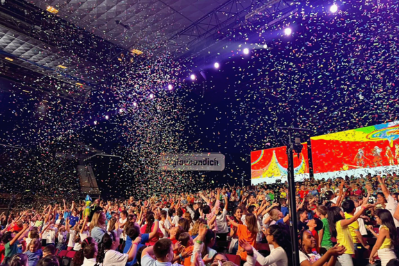 Volksschulkinder mit bunten T-Shirts in einer großen Halle, weiße Spots in violett-blauem Licht und Konfettiregen.