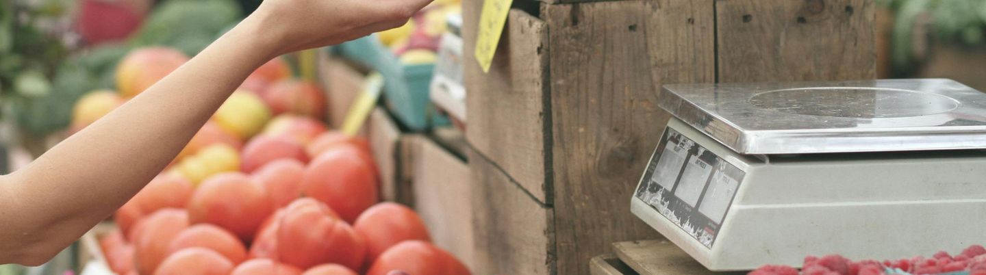 A person's hand receiving an apple from another's over a market stall filled with fresh produce, with a weighing scale on the side.