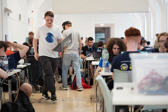Raum mit Hacker_innen sitzend, einer stehend mit Laptop in der Hand