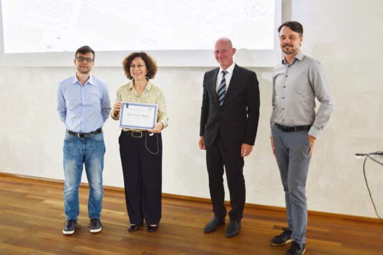 Gruppe von vier Menschen vor einer Leinwand, die zweite von links ist eine Frau, die eine Urkunde präsentiert.