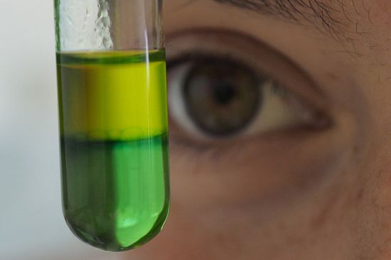 Close-up of a glass tube with two differently coloured separated liquids (green and yellow), behind it part of a face with an eye 