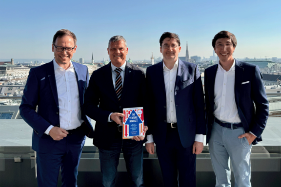 Gruppenbild mit vier Personen auf einer Dachterrasse. Im Hintergrund blauer Himmel und die Dächer Wiens.