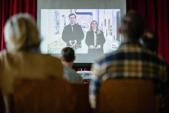 Personen sitzen in einem Raum und schauen ein Video an. 