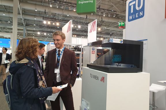 On the left a woman talking to Christopher Lackner (TU Wien) abiut NGSolve, on the right the hologram pyramid shows a simulation of the softwares functionality.
