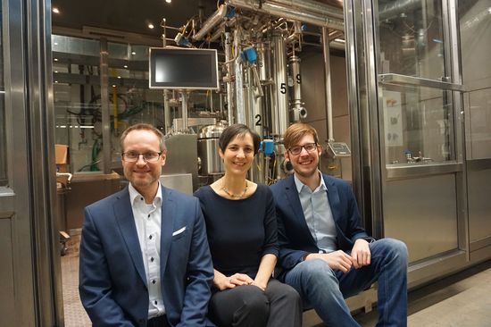 Drei Personen im Labor, dahinter eine Anlage mit Röhren und einem Bildschirm