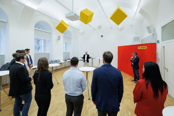 Foto von der Vergabe der PALFINGER Elevate Stipendien: Man sieht rechts ein großes rotes Rollup von Palfinger, davor steht Rektor Jens Schneider. Daneben steht Philipp Smole von PALFINGER und begrüßt die Stipendiat_innen, die mit dem Rücken zur Kamera stehen. 