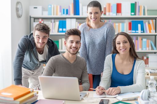 junge Menschen sitzend an und stehend hinter Tisch, auf dem Tisch Laptop und Bücher, im Hintergrund Bücherregal