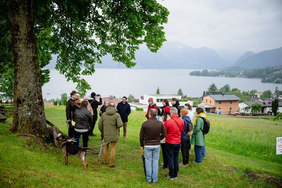 eine Gruppe Menschen steht im Kreis auf einer Wiese, links ein Baum, im Hintergrund ein See und Berge