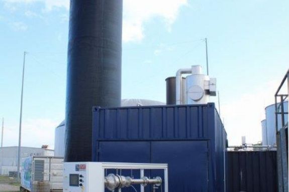 Neuartiger Kurzzeitkontaktor der TU Wien im Größenvergleich zur heute üblichen Entschwefelungstechnik in weißem Container vor altem, ein Vielfaches größerem Anlagentyp