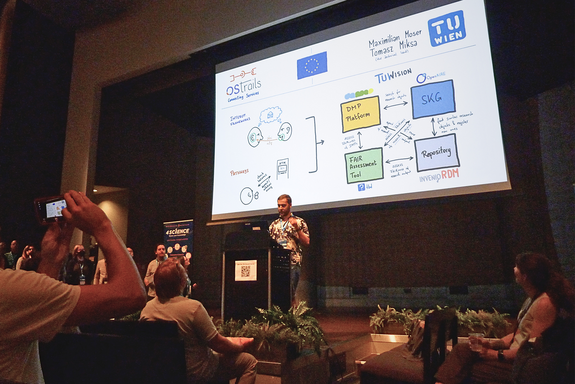 The photo shows an audience, a large screen with a presentation slide, and a person standing in front of the screen at a speaker's podium.