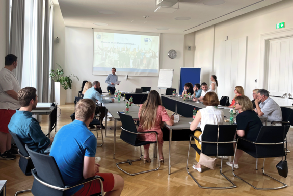 Seminar participants in a room with parquet floor and screen in the background. They sit together in a U-shape.