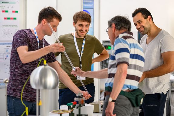 Drei Wissenschaftler zeigen einem Besucher, wie eine Kugel in einem Rohr abgebremst wird.