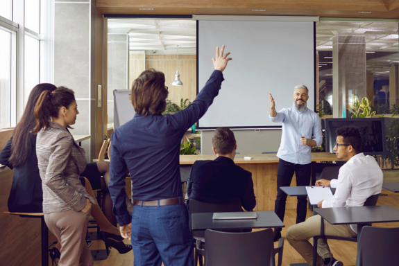 Moderner Seminarraum mit Vortragendem vor Screen. Von hinten sichtbar Studierende in Businesskleidung, von denen einer aufzeigt.