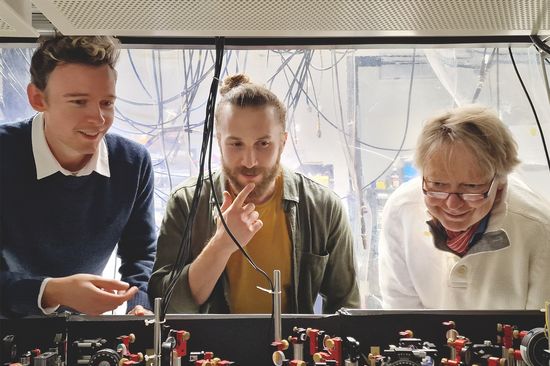 Drei Personen im Labor, vor ihnen eine optische Bank mit Spiegeln und Linsen