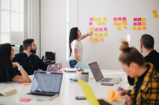 The photo shows people of different genders and skin colors in an office. They are sitting around a table with open laptops and working on something. A woman is standing at the front of the whiteboard with colorful Post-Its on it and pointing at a sticky note.