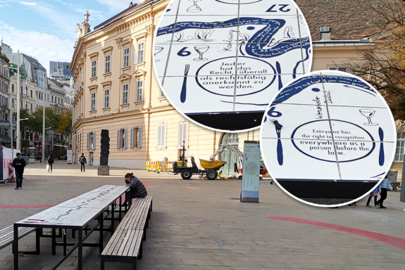 Überdimensionale runde Plaketten mit Artikel 6 der Menschrechte rechts hinten, vorne links eine Tisch auf dessen Platte die Menschenrechte notiert sind. Szene: Platz der Menschenrechte, Mariahilferstraße, Wien, Übergangszeit, Sonnenlicht.
