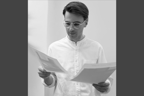 Schwarz-weiß-Brustbild mit grauen Balken einer Person, die ein Hemd mit Stehkragen trägt und in beiden Händen ein Blatt Papier hält.