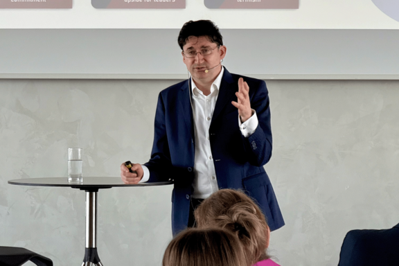 Person mit dunklen, gelockten Haaren während eines Vortrags. Er trägt ein weißes Hemd und dunkelblaues Sakko und lehnt leicht an einem Stehtisch