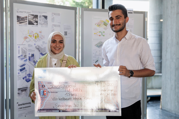 Zwei Personen halten einen überdimensionalen Scheck auf Brusthöhe hoch. Im Hintergrund eine Wand mit Planungsentwürfen.
