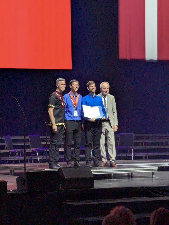 Überreichung der silbernen Medaille samt Urkunde durch Jurymitglieder. In der Mitte: Chorleiter Andreas Ipp mit Medaille umgehängt (2. von links) und sein Stellvertreter Alexander Pacha mit Urkunde (3. von links). Daneben stehen 2 Männer von der Jury. Sie stehen alle auf der Bühne. Im Hintergrund sieht man die österreichische Fahne.